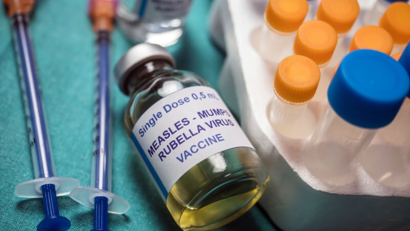 A vial containing the Measles, Mumps, Rubella (MMR) vaccine is photographed with syringes and other vials. 