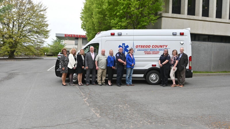 11 people stand in front of an Otsego County Emergency Medical Services vehicle. Text on the side of the vehicle reads "Proudly Serving Otsego County." The vehicle is parked in front of the emergency department entrance at Bassett Medical Center.