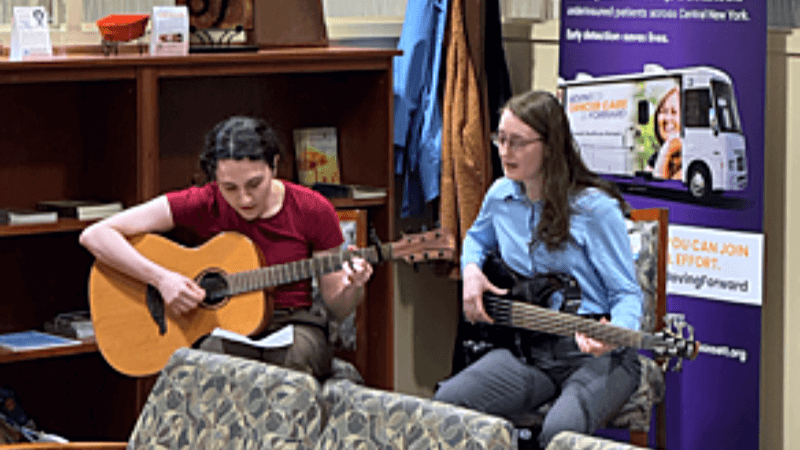 Two people are seated in a hospital waiting room. They are each strumming a guitar.