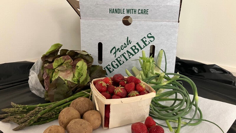 An open box that says "Fresh Vegetables" on the side is on a table. In front of the box are various produce items, including a pint of strawberries and five potatoes.
