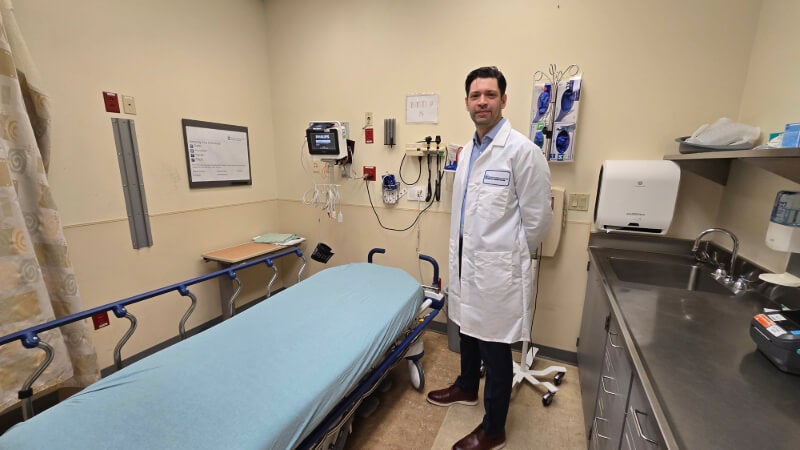 A man in a white doctors coat stands next in an Emergency Room setting. A counter and a patient bed is seen. 
