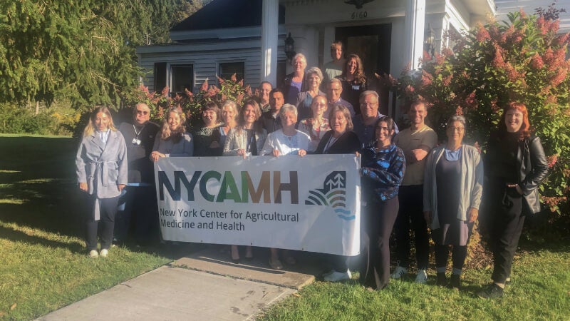 A group of people stands in front of a building with a large banner that reads NYCAMH New York Center for Agricultural Medicine and Health