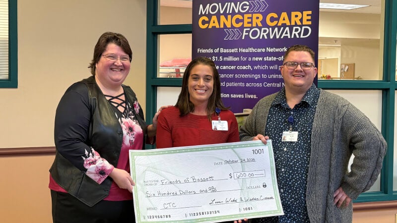 Three people stand with a large ceremonial check for 600 dollars. They are in front of a banner that reads "Moving Cancer Care Forward" 