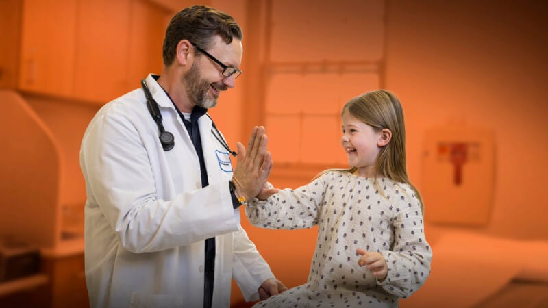 A man in a white doctor's coat with a stethoscope around his neck gives a high five to a young girl in an exam room. 