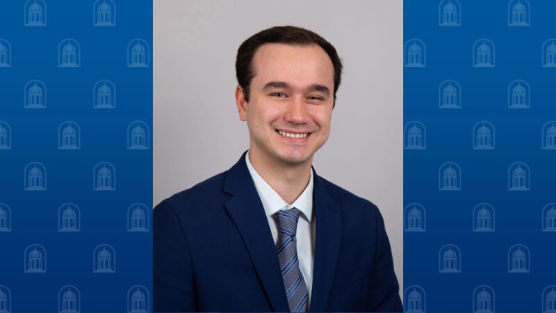 A man wearing a suit and tie is photographed from the shoulders up. He has short black hair and he is smiling. 