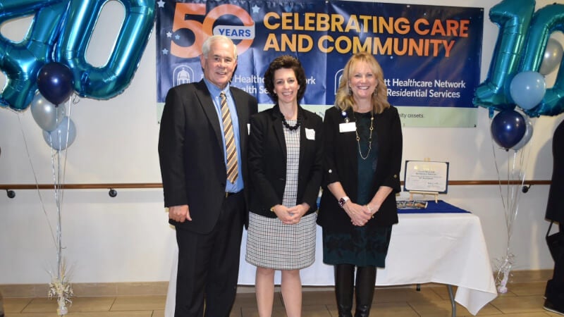 Three people dressed in professional attire stand for a photo. Behind them is a banner that reads "50 years: Celebrating Care and Community" and balloons in the shape of the numbers 40 and 10.