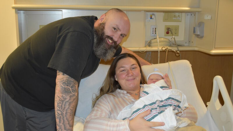 A man stands next to a woman in a hospital bed. The woman is holding a newborn baby.