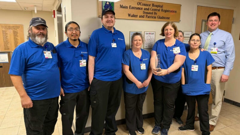 Eight people wearing blue shirts are photographed in a hospital setting. One of them is holding an award made of glass. 