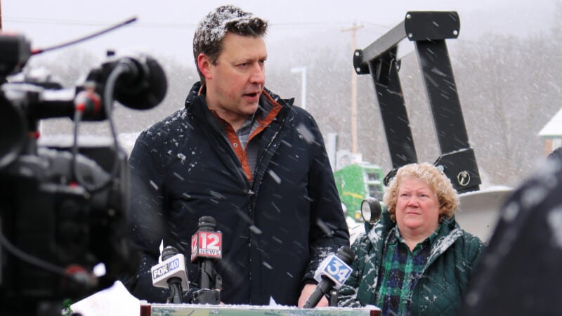 A man, Congressman Josh Riley, speaks at a podium outside. There is a video camera recording him and a woman standing behind him.