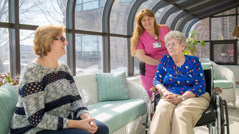 A CNA in pink scrubs stands with two elderly ladies. The three people are seated in a sunroom area. 