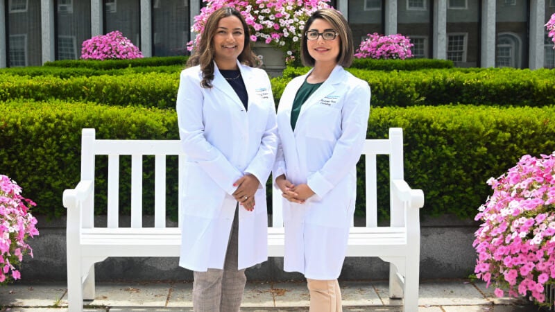 Two doctors in white coats, Dr. Roudy Kiani and Dr. Anjali Prakash, are photographed outside in front of hedges, flowers, and a white park bench. 