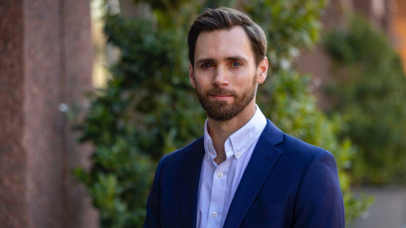 A man, Dr. Tyler Malone, is photographed from the shoulders up in front of some greenery. He has short hair, a beard, and is wearing a blazer and button down shirt. 