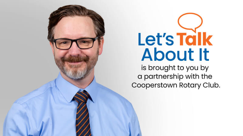 A man, Dr. Brandon Greene, is photographed. He is wearing a button down shirt, tie, and glasses. Text on the image reads "Let's Talk About It is brought to you by a partnership with the Cooperstown Rotary Club" 