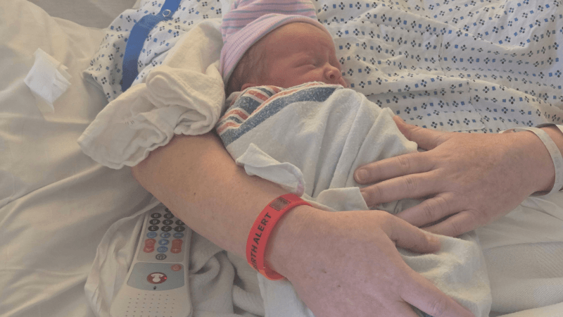 A newborn baby is held up her mom. The mom's face is not seen, but her wrist has a red health alert wristband on it.