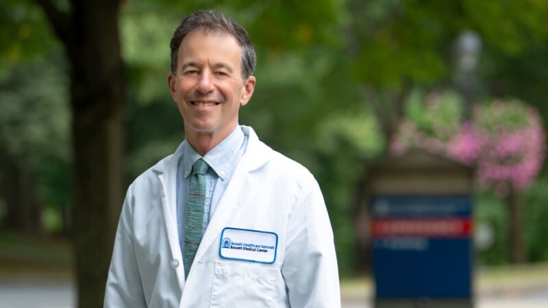 A man in a white doctor's coat, Dr. Henry Weil, stands outside in front of a Bassett Medical Center sign.