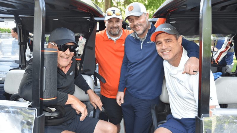  Four people are photographed. Two of them are seated in golf carts. The other two are standing between the golf carts.