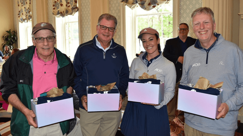 Four golfers stand together for a photo. They are each holding a gift.