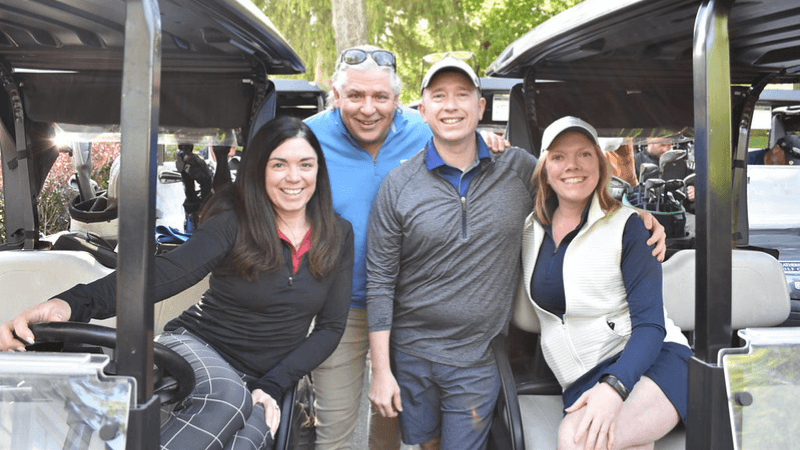 Four people are photographed. Two of them are seated in golf carts. The other two are standing between the golf carts.