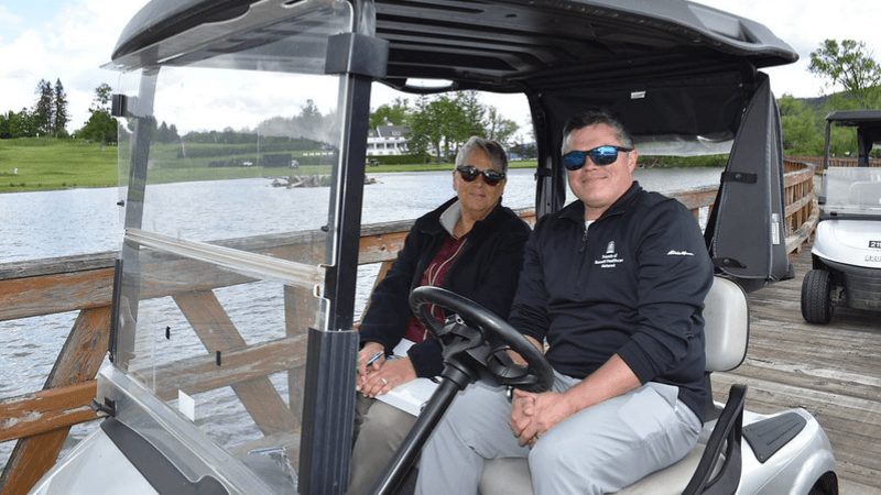 Two people are photographed seated in a golf cart. The beauty of the Otesaga Hotel’s Leatherstocking Golf Course. can be seen behind them.