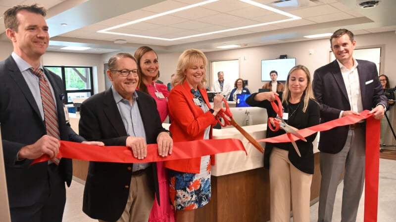 Six people hold up a red ribbon inside a medical building. Two of the people are cutting the ribbon with oversized ceremonial scissors.
