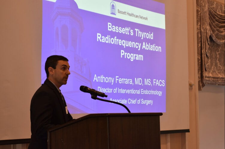 A person stands at a podium. A presentation side is projected on a screen behind them.
