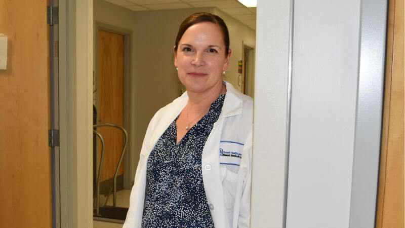 A woman in a white doctor's coat stands in a hallway.