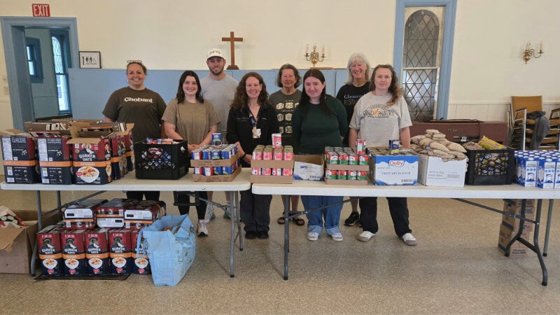 A group of eight people stand behind a table to smile for a photo. The table is covered in boxes of various non-perishable food items. 