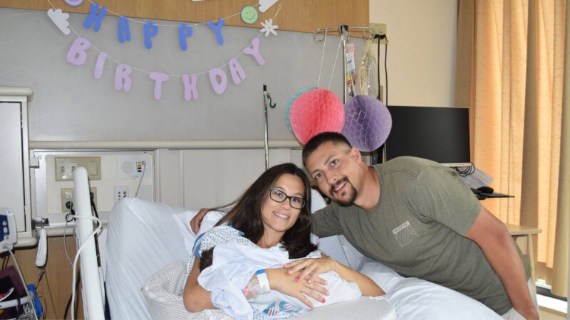 A new mother holds her baby in her hospital bed. A man stands leans over her and places his arm around her shoulders. A banner on the wall behind the family reads "Happy Birthday"