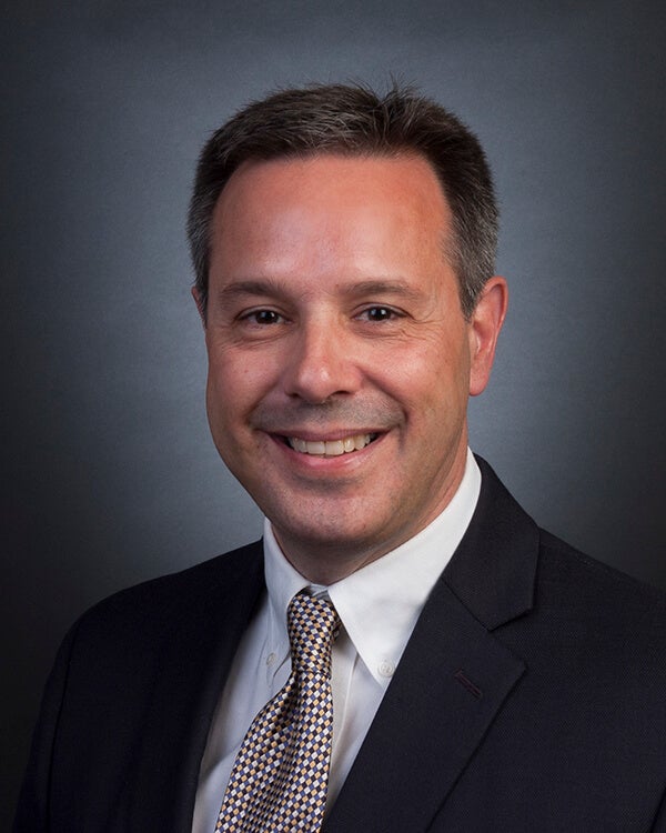 A man is photographed from the shoulders up. He is wearing professional attire.