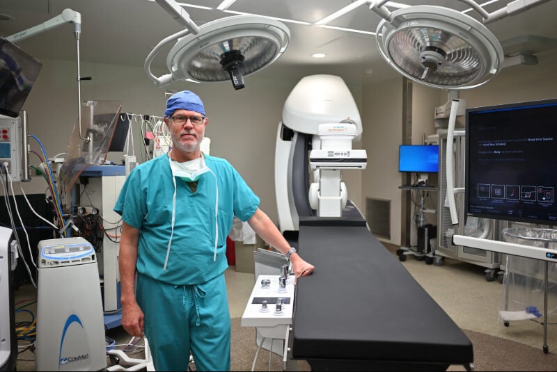 A man stands next to an operating table in Bassett Medical Center's hybrid operating room. He is wearing scrubs and a surgical cap.