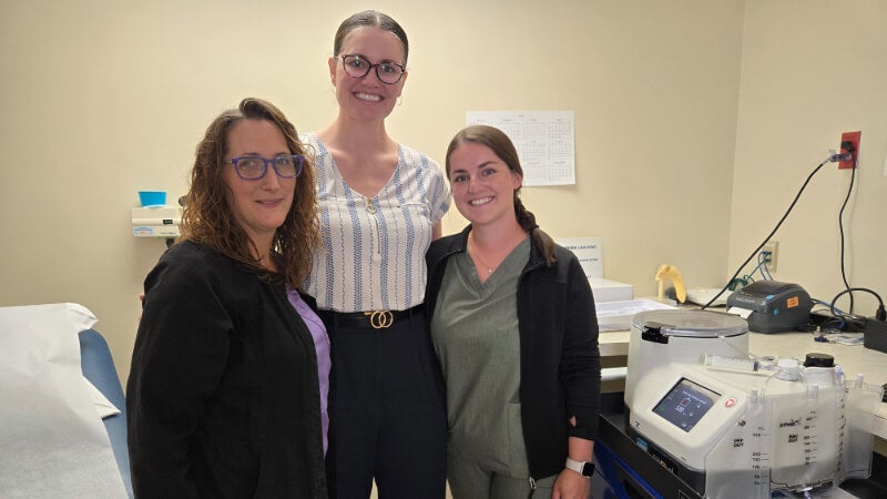 Three women are photographed standing next to a centrifuge.