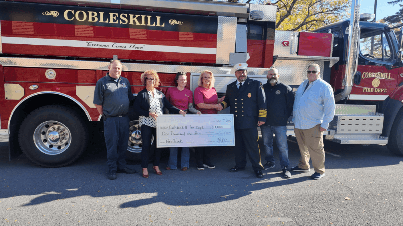 Seven people are photographed in front of a fire engine. Two of them are shaking hands. Another two are holding up a large ceremonial check. The fire engine behind them has the Cobleskill Fire Department logo on it. 