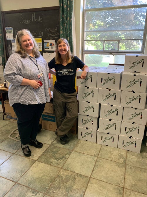 Two women stand next to boxes of fresh vegetables.