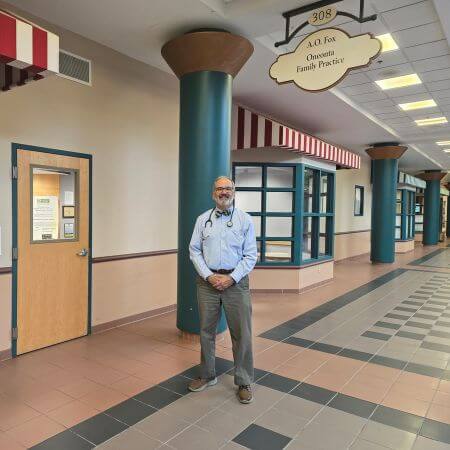 A man is standing inside FoxCare Center. A sign that reads "A.O. Fox Oneonta Family Practice" hangs above his head. 