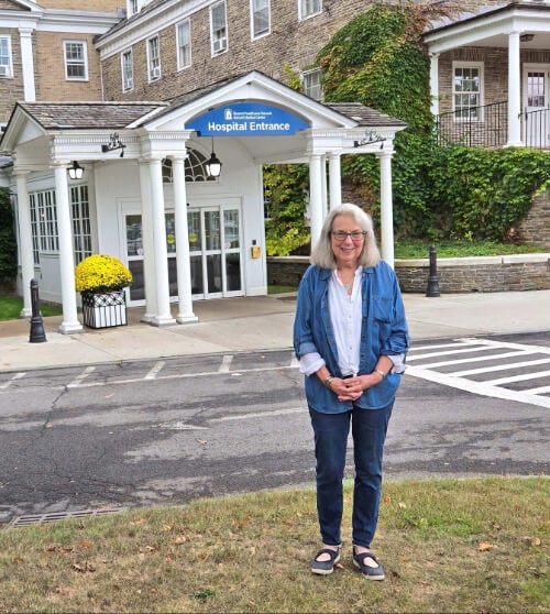 A woman, Georgia Meeter, stands outside Bassett Medical Center. She is wearing jeans, a white button down shirt, and a denim overshirt.