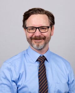 A man wearing a professional-looking tie and button down shirt is photographed. He has brown hair, a grey beard, and glasses.