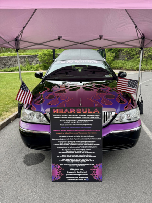 A hearse with a flame wrap in the colors black, purple, orange, and lavender is parked under a tent. A sign in front of it lists its name, Hearsula, and purpose.