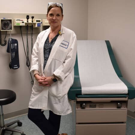 A woman in a white doctor's coat stands in a medical exam room. 
