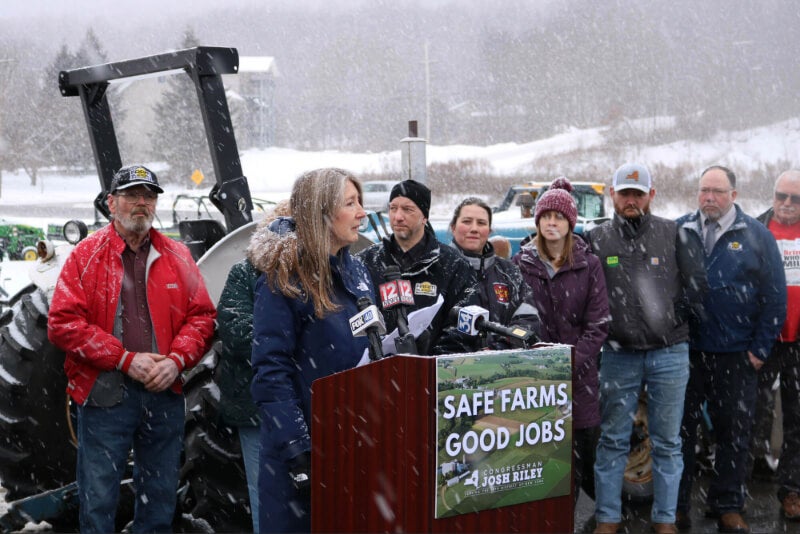 A woman, Julie Sorensen, stands at a podium with farmers and other officials behind her. The podium reads "Safe Farms. Good Jobs. Congressman Josh Riley."