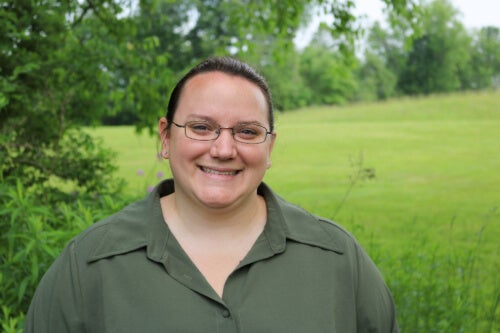 A woman wearing glasses and a button down shirt is standing outside. She is smiling.