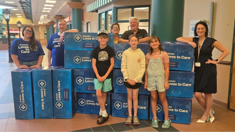 Eight people stand for a photo with over a dozen boxes. The boxes are branded with "Subaru Loves to Care" designwork. 