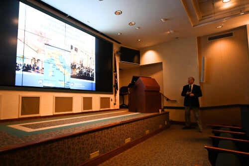 A man stands in front of a podium, delivering a lecture. He is looking at a large screen on which is presentation can be seen.