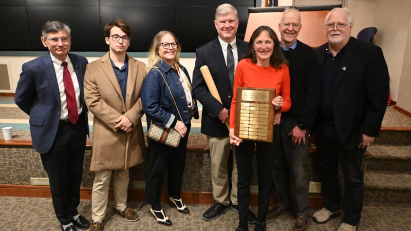 Seven people stand for a photo. One of them is holding a commemorative bat and another is holding a plaque.  