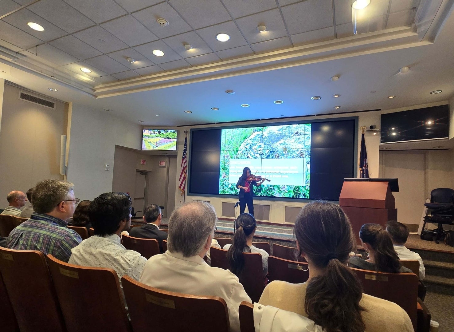 A woman plays a viola next to a podium. Audience members look on.