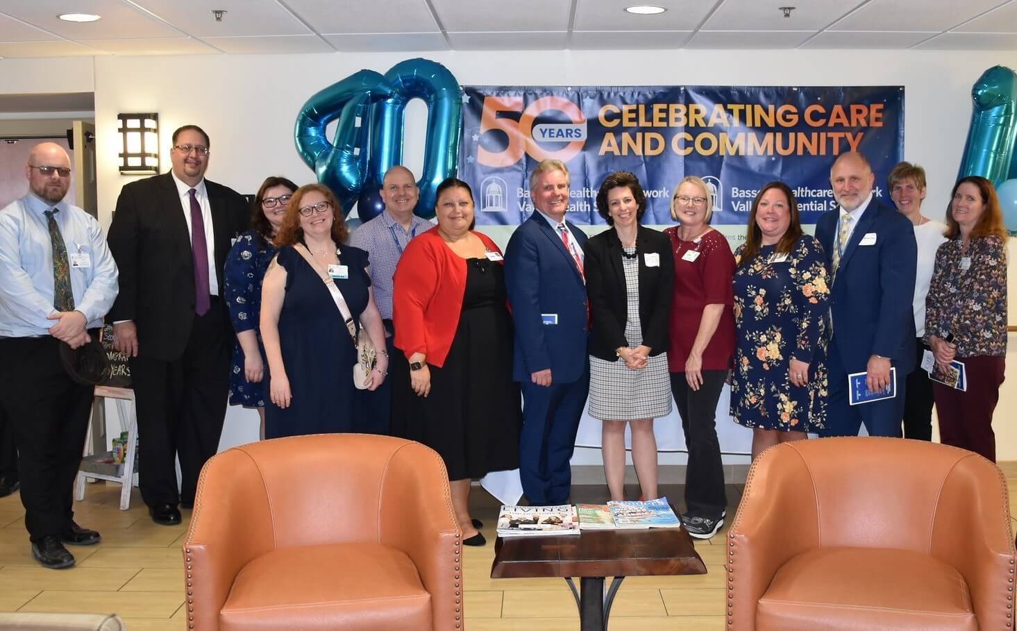 Thirteen people  stand for a photo. Behind them is a banner that reads "50 years: Celebrating Care and Community" and balloons in the shape of the numbers 40 and 10.