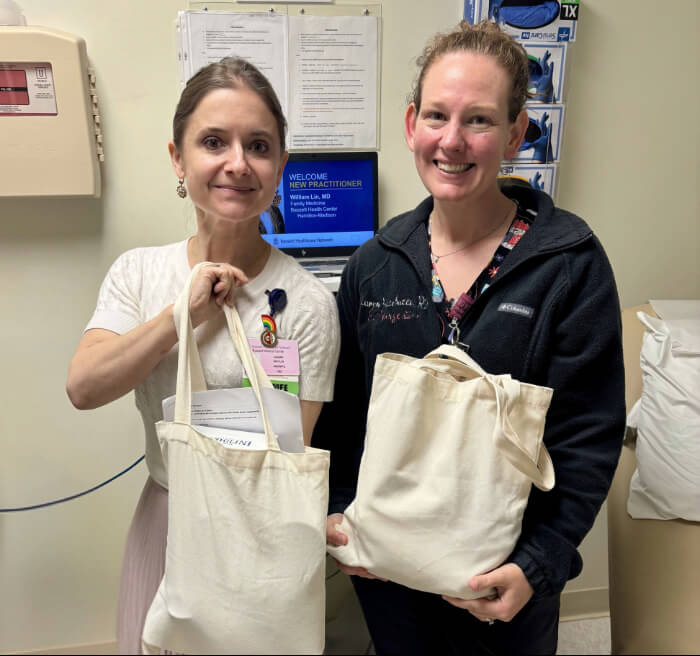 Two women are photographed in a hospital setting. They are each holding a canvas tote bag